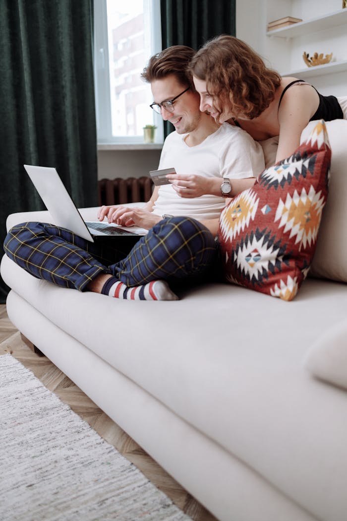 Happy couple sitting on a couch, using a laptop for online shopping with a credit card in hand.